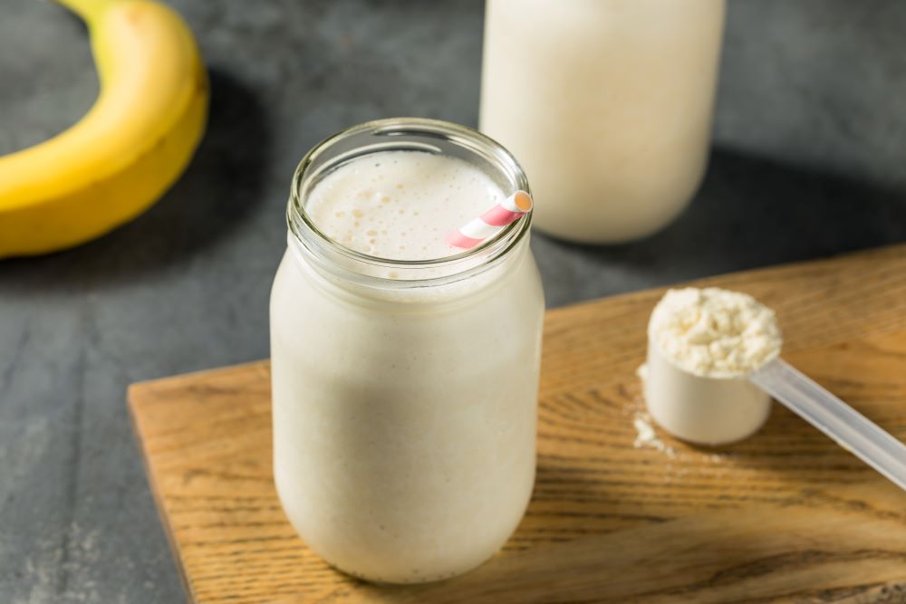 Whey protein shake in a glass jar with a straw, with scoop of whey protein adjacent and a banana in the background