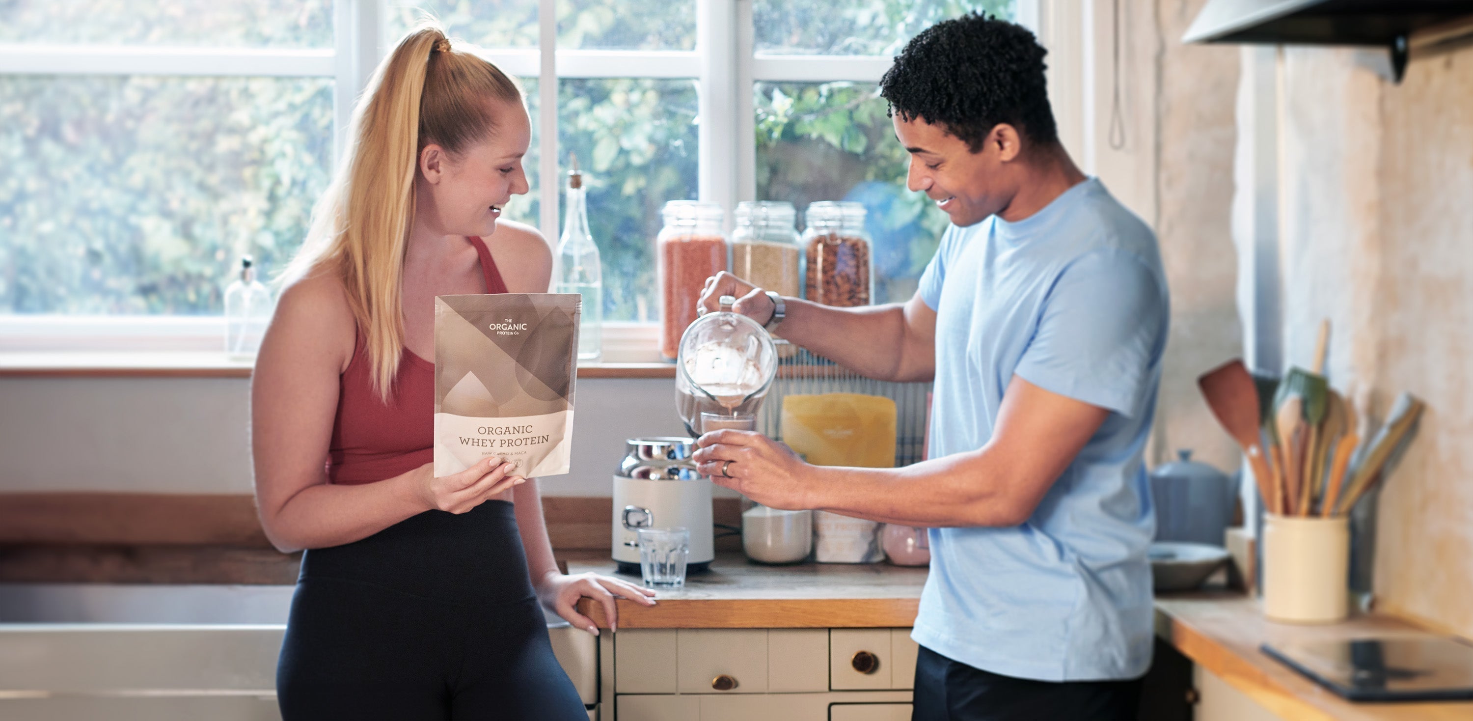 A man and woman are standing in a kitchen pouring an organic whey protein shake into a glass from a blender