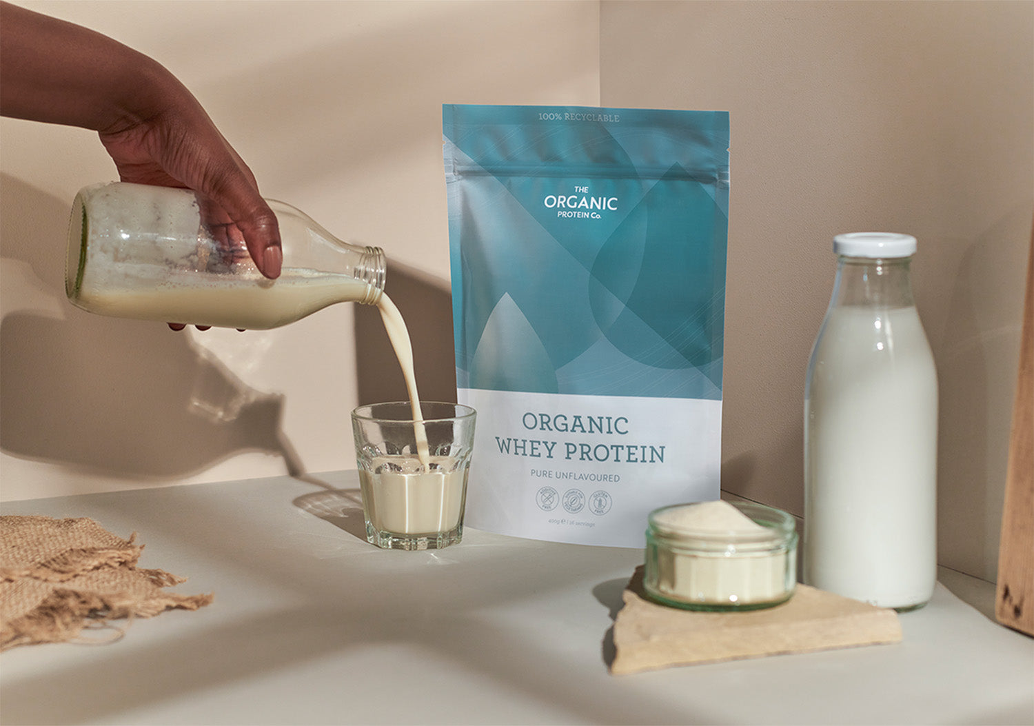 Person pouring unflavoured organic whey protein powder into glass, next to a pouch of whey protein and a bottle of milk